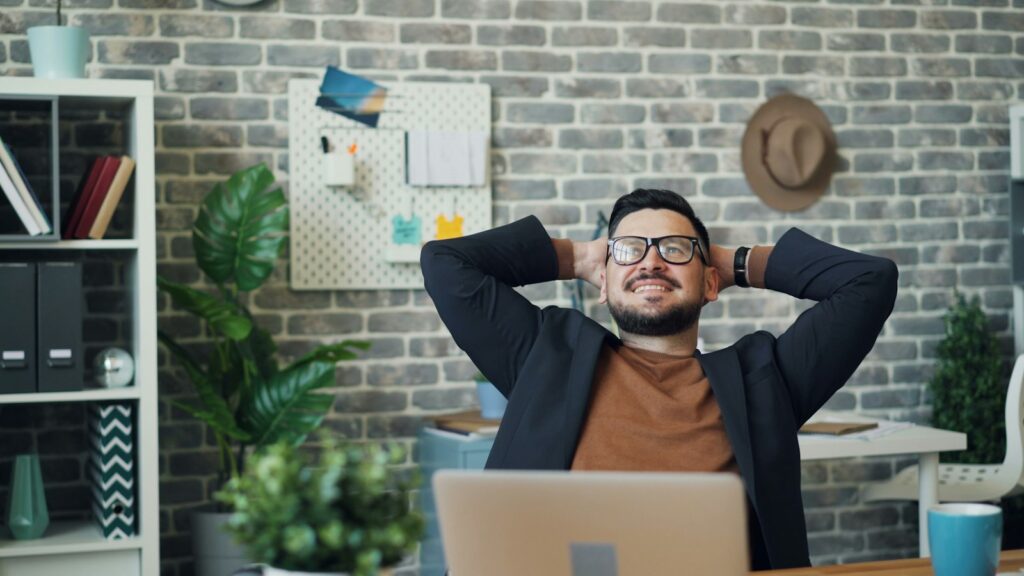 Mann sitzt am Schreibtisch im Büro und streckt sich mit den Händen hinter dem Kopf