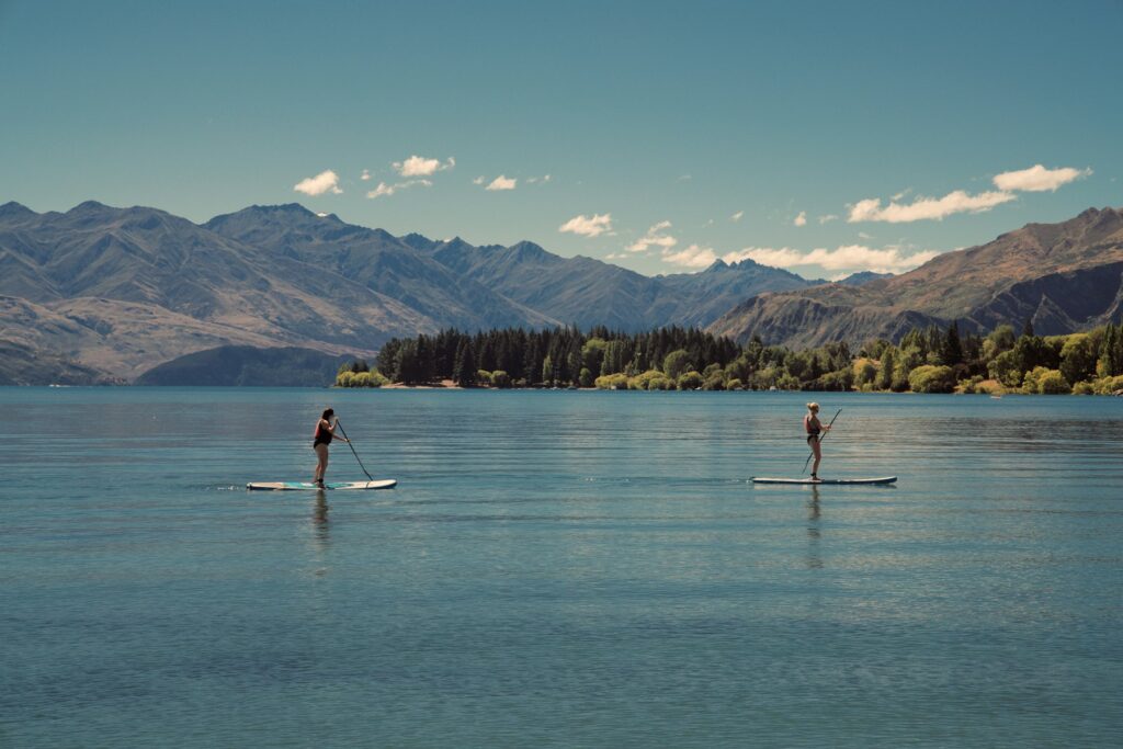 Zwei Personen paddeln stehend auf SUP-Boards über einen See vor Bergen